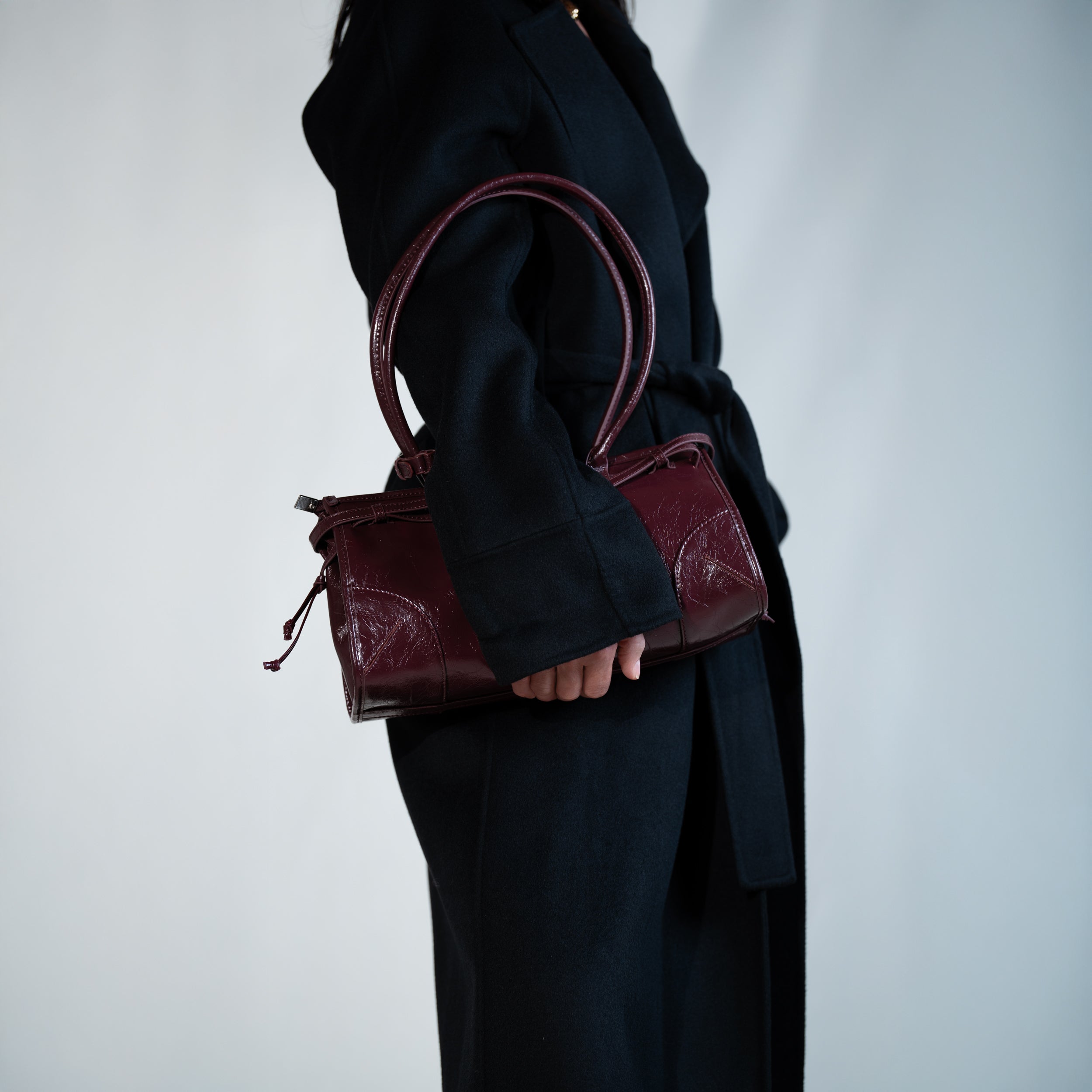 sac à main forme baguette femme en cuir verni bordeaux vintage porté sous le bras sur un manteau noir de côté