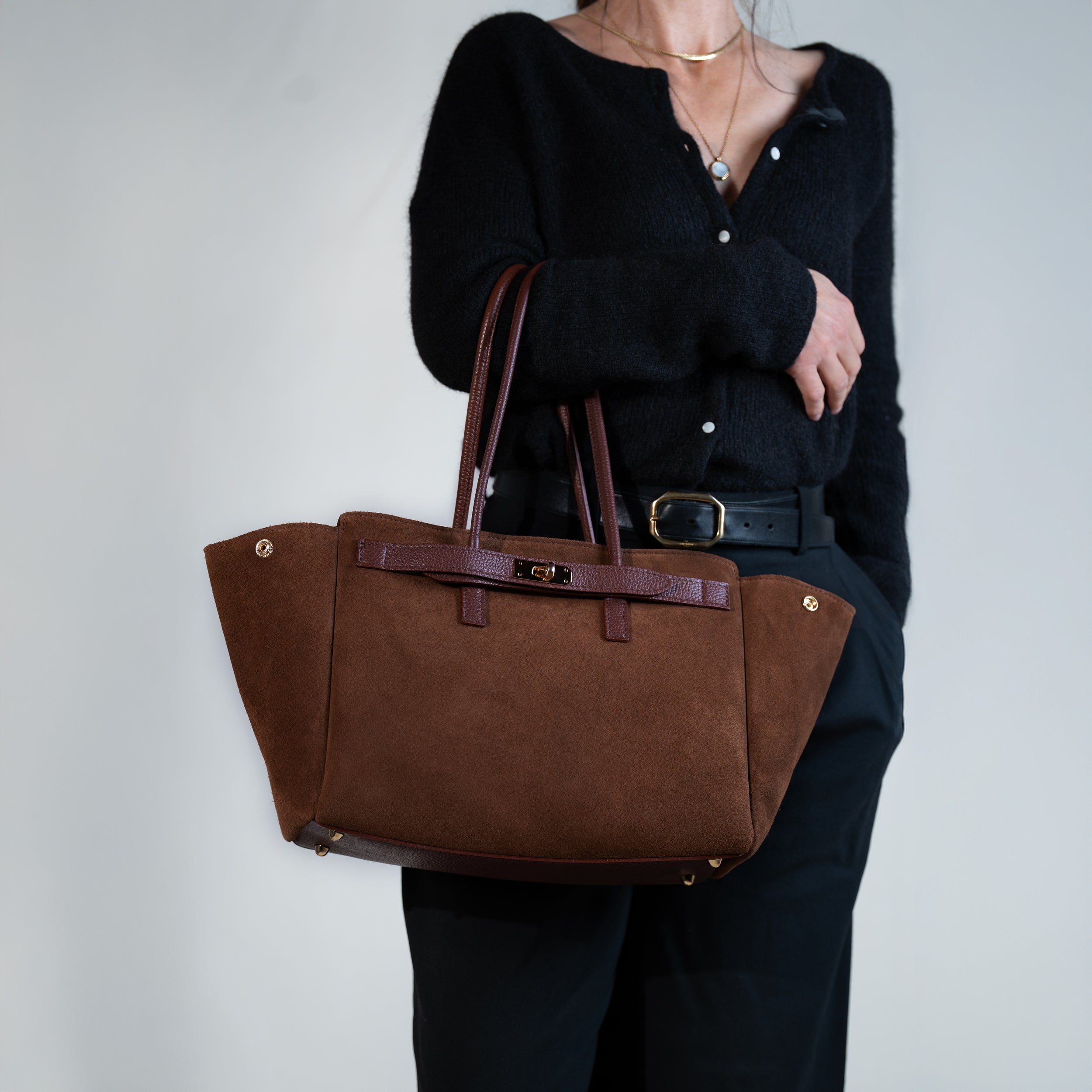 sac cabas femme en cuir suédé ou daim camel en forme trapèze avec fermoir tourniquet porté au bras sur une tenue avec un pull noir, une ceinture noir et un pantalon tailleur noir