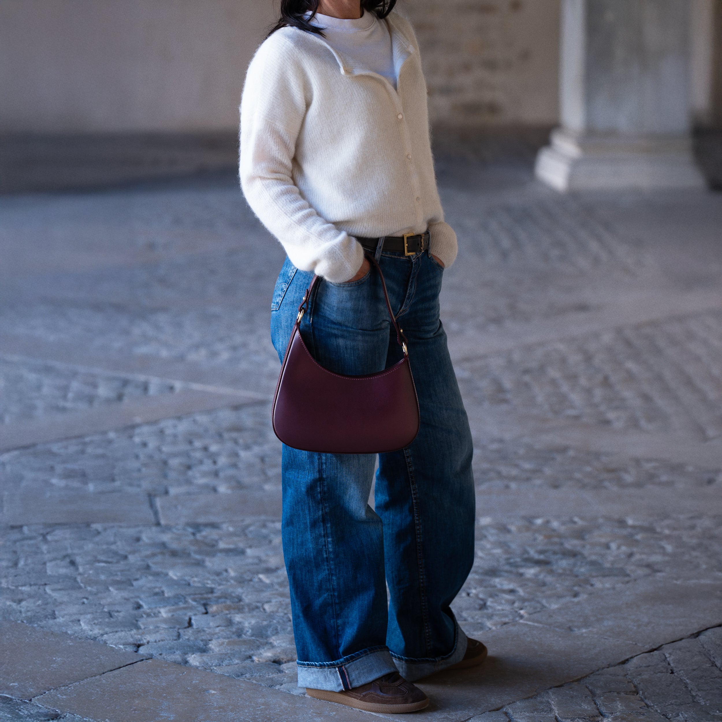 sac à main forme hobo demi lune asymétrique pour femme en cuir lisse bordeaux porté à la main sur jeans et pull blanc lifestyle