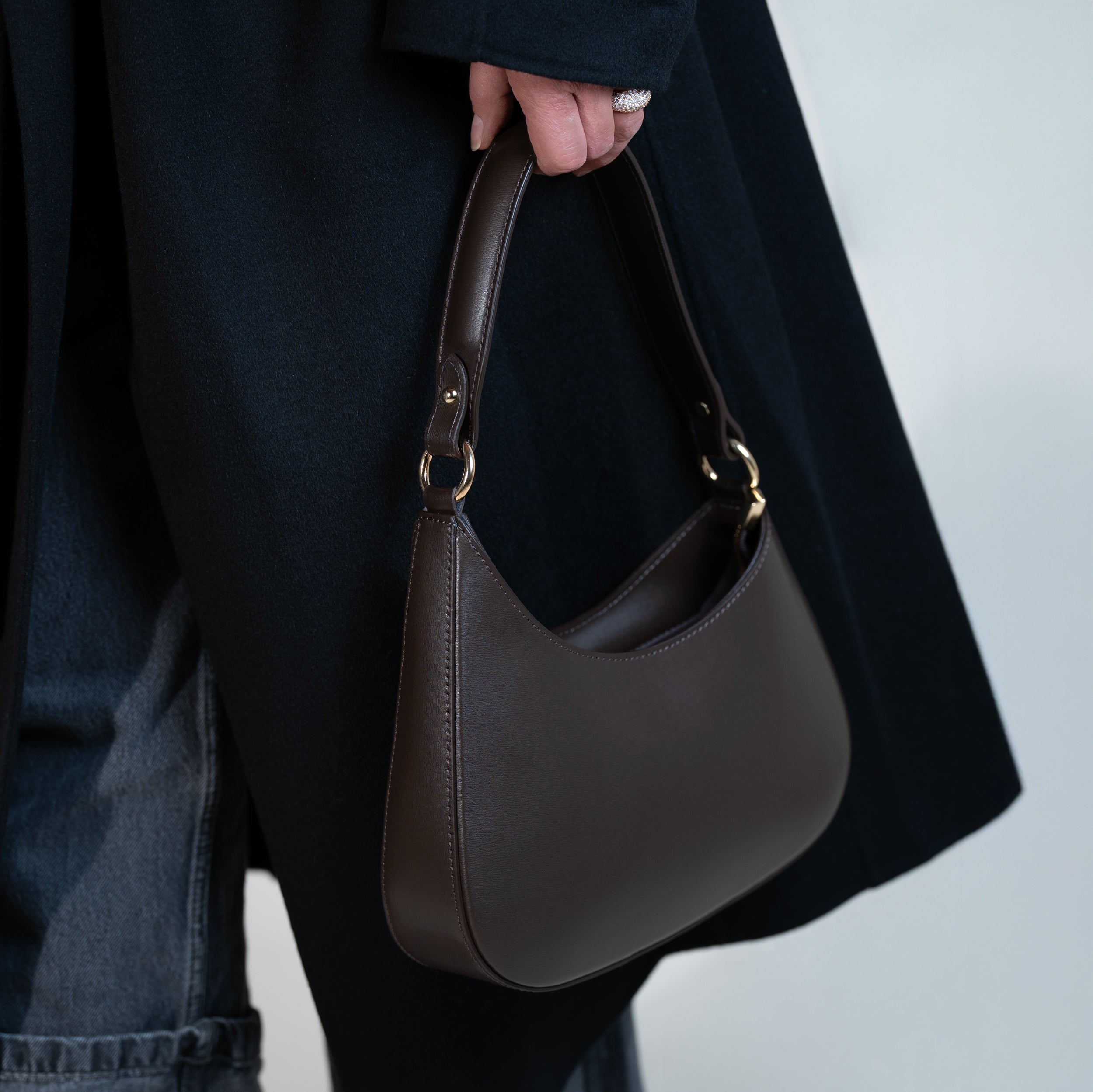 sac à main forme hobo demi lune asymétrique pour femme en cuir lisse marron porté à la main sur un manteau noir et un jean noir