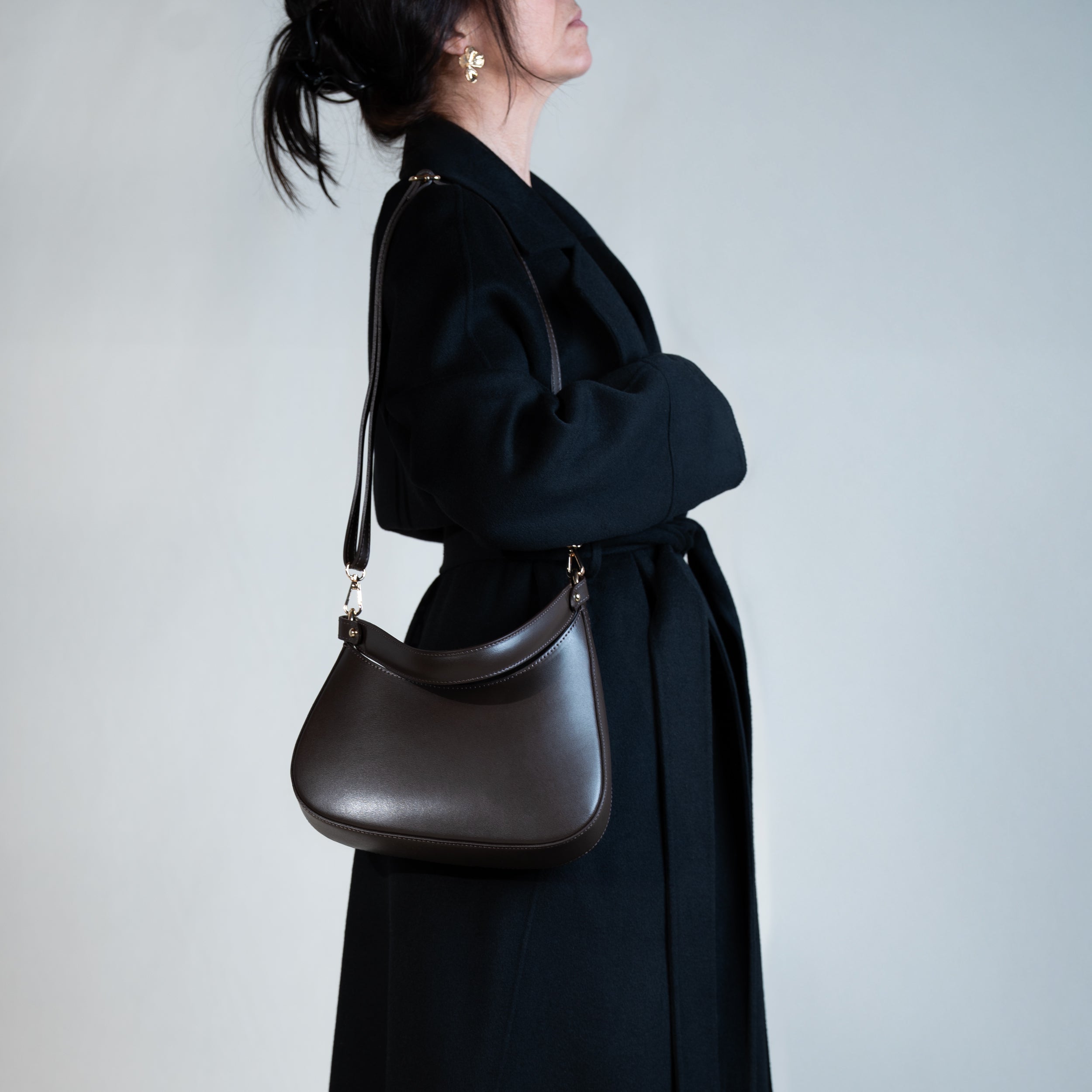 sac à main forme hobo demi lune asymétrique pour femme en cuir lisse marron porté en bandoulière avec manteau noir et boucles d'oreilles fleur