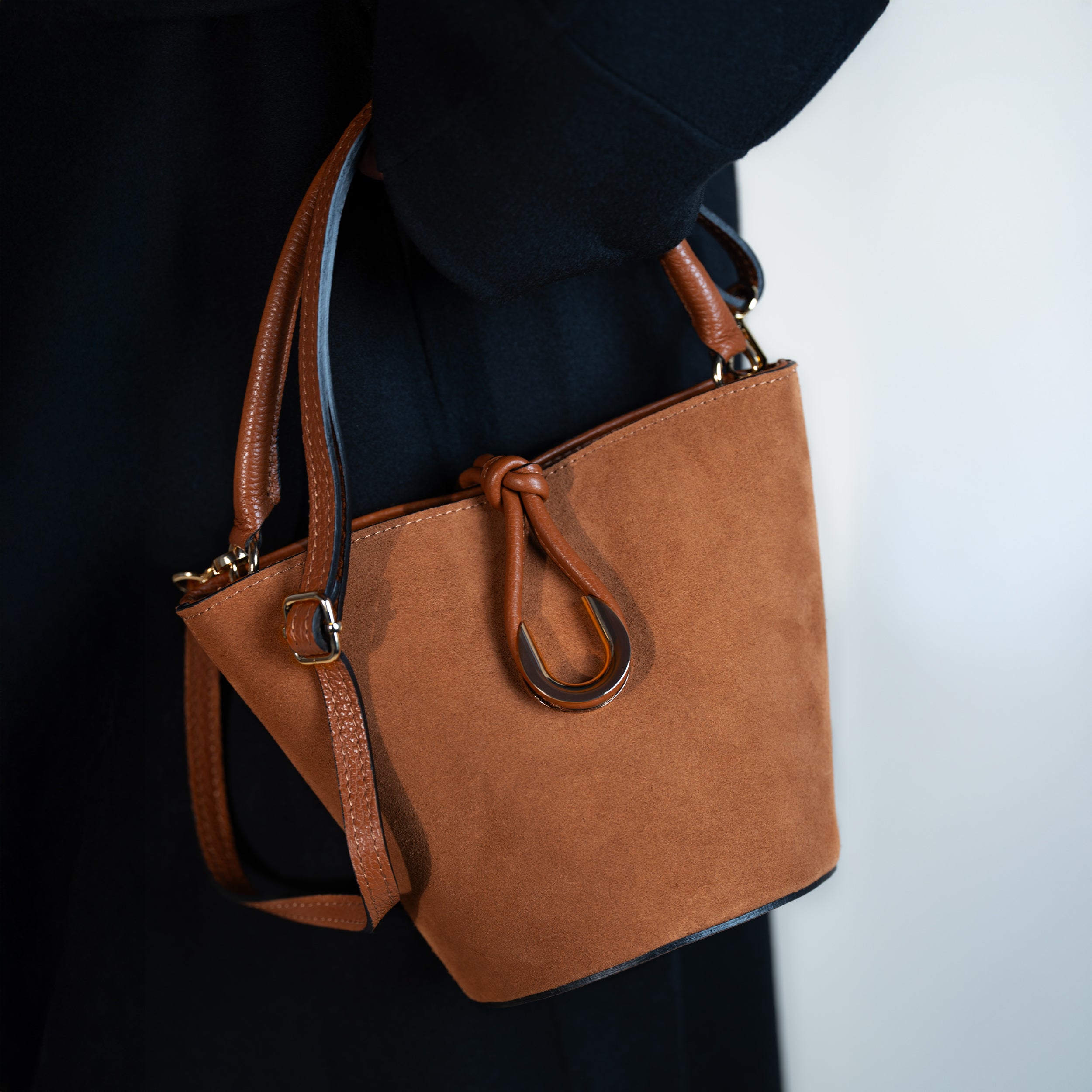sac à main femme en cuir suédé ou daim camel en forme seau porté à la main avec une bandoulière et un fermoir doré original sur un manteau noir