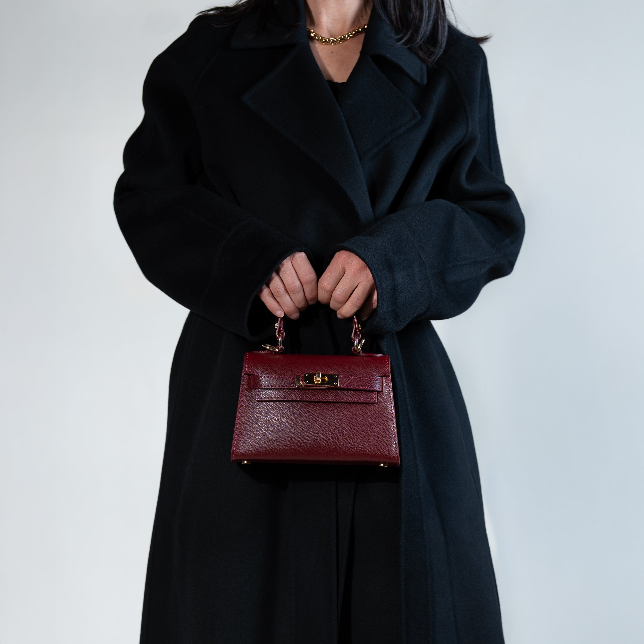 sac à main femme en cuir saffiano bordeau et bijouterie dorée dans une forme top handle porté à la main avec un manteau noir 