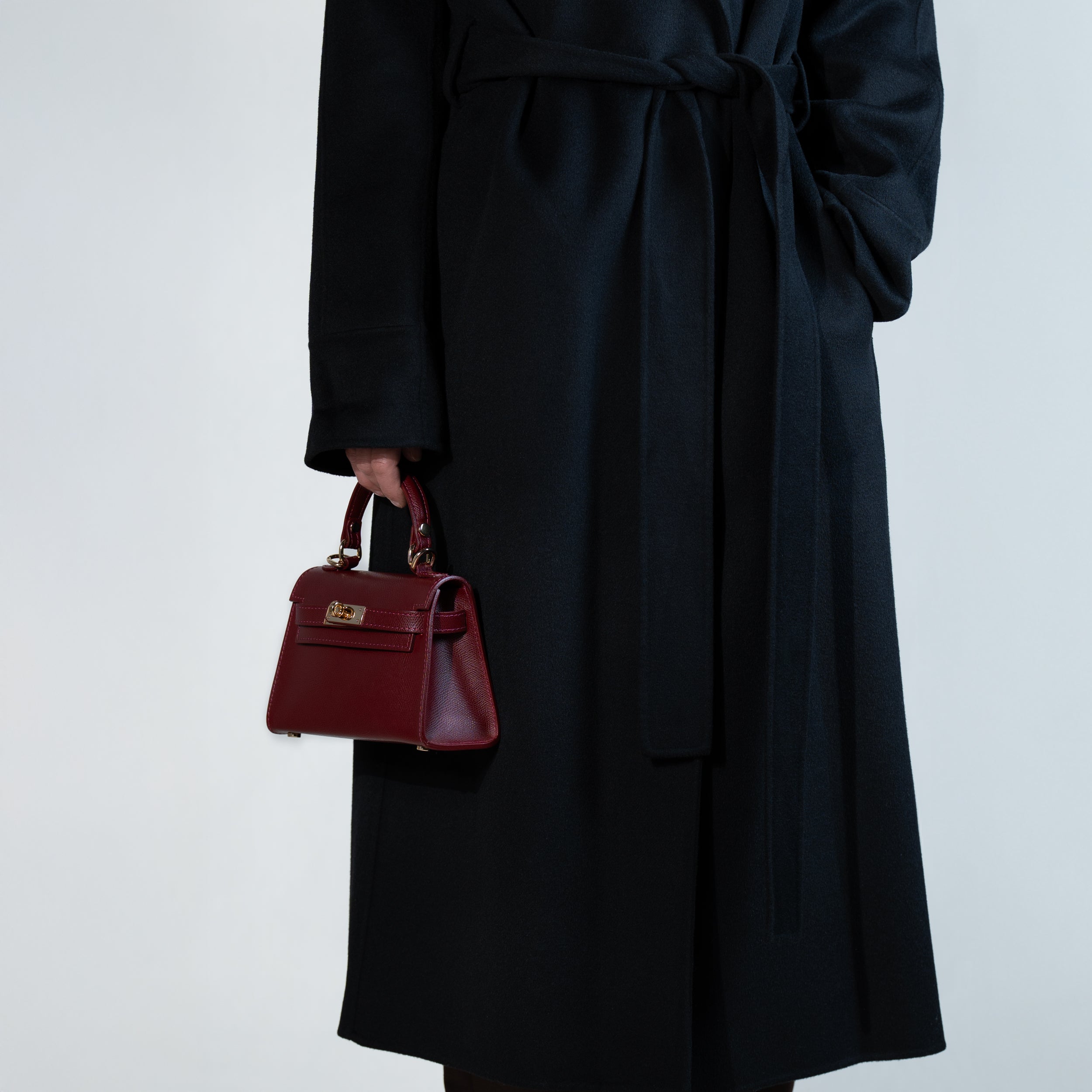 sac à main femme en cuir saffiano top handle porté à la main sur un manteau noir  avec fermoir tourniquet et bijouterie doré dans un cuir de qualité bordeaux 