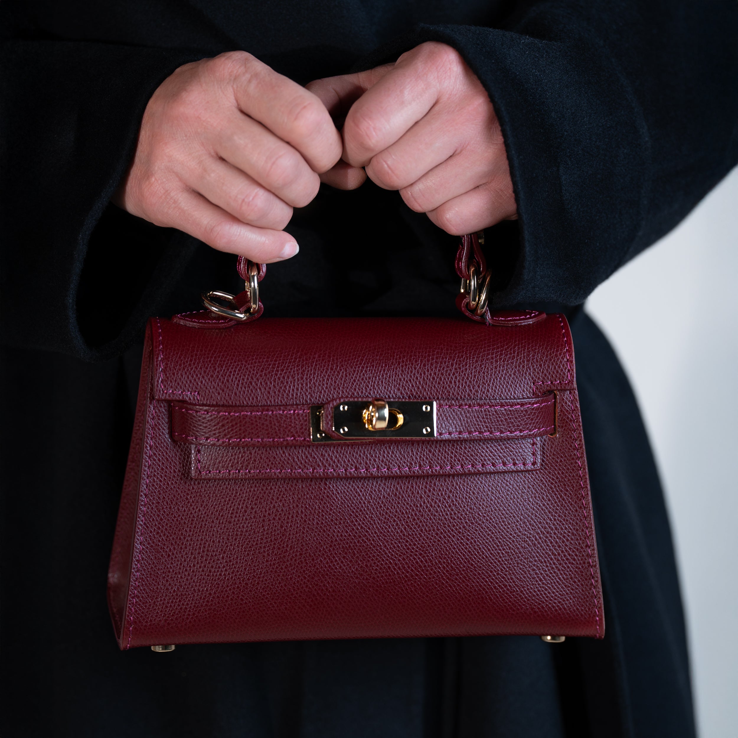 sac à main femme en cuir saffiano top handle avec fermoir tourniquet et bijouterie doré dans un cuir de qualité bordeaux porté à la main sur un manteau noir 