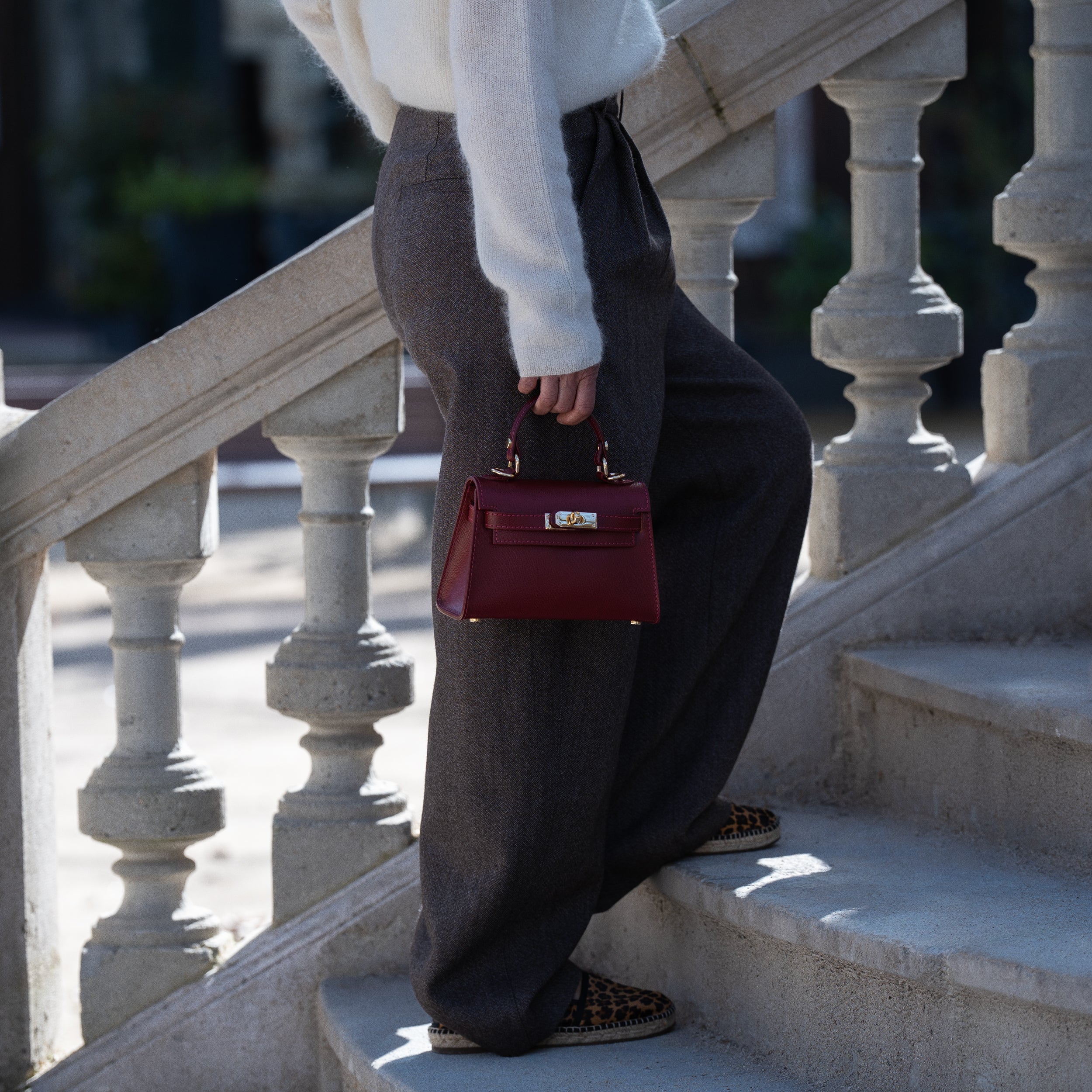 sac à main top handle pour femme en cuir saffiano bordeaux porté à la main sur pantalon marron et pull blanc